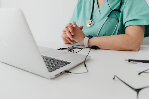 Medical practitioner in scrub suit using a laptop for remote consultation and documentation.