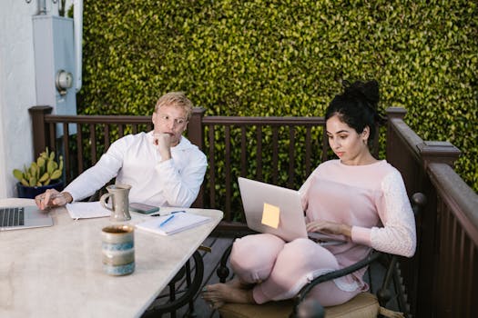 Two adults working remotely on laptops outdoors, discussing and collaborating.