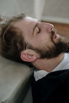 Close-up side view of a bearded man resting with eyes closed, in a calm indoor setting.